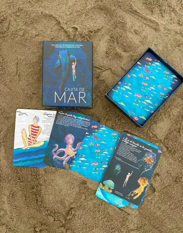 La Cajita de Mar abierta en la arena, junto a cuatro de sus cartas ilustradas.